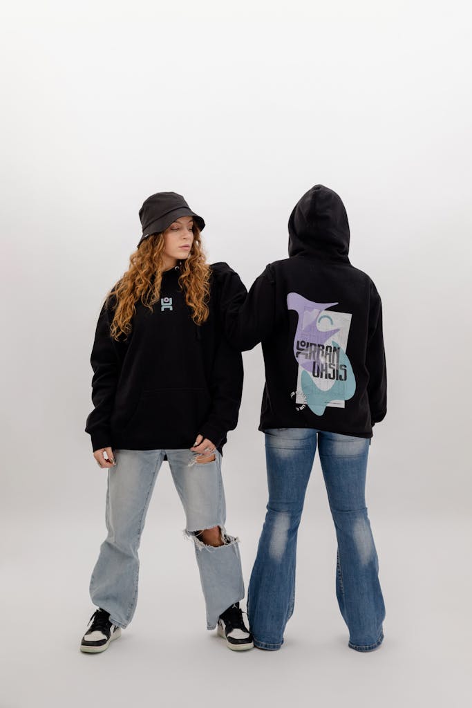 Two young adults in stylish urban outfits with hoodies and jeans, standing indoors.