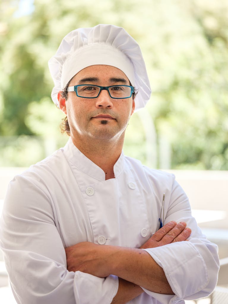 Professional male chef wearing glasses and toque blanche, standing confidently with crossed arms.