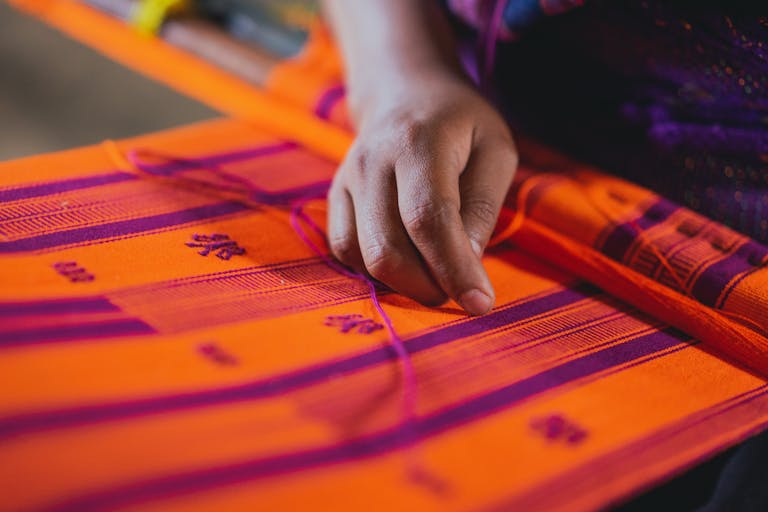 Close-up of a hand weaving a vibrant orange and purple textile, showcasing traditional craftsmanship.